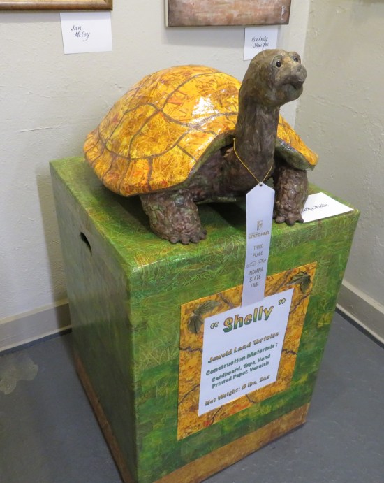 A tortoise sculpture on a tall green dais with a very large name tag.