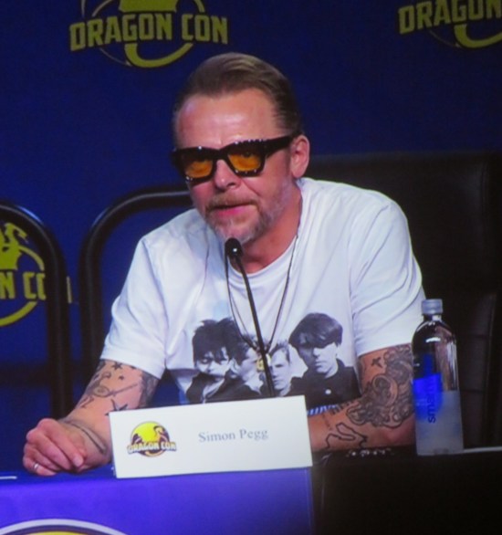 Simon Pegg on big-screen TV, sitting at Q&A table with name card and mic.