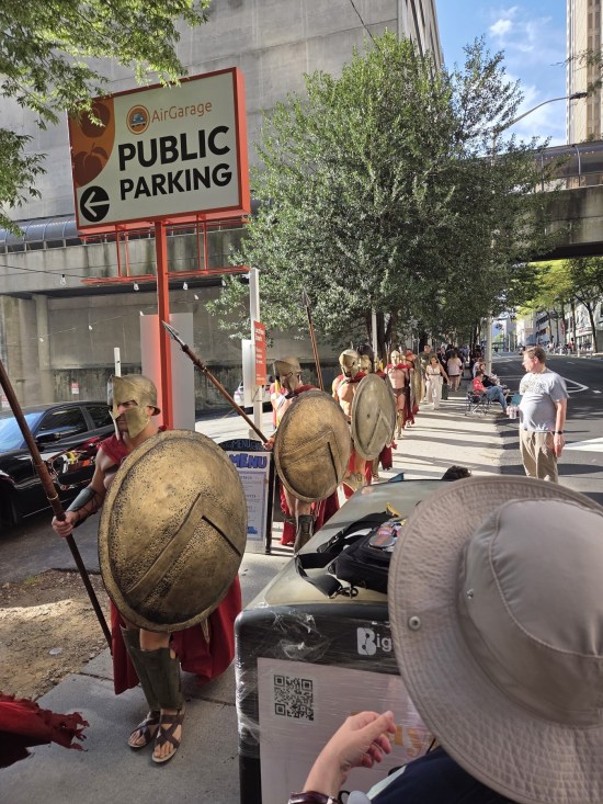 Spartan costumes from Frank Miller's "300", marching down a city street next to a public parking lot.