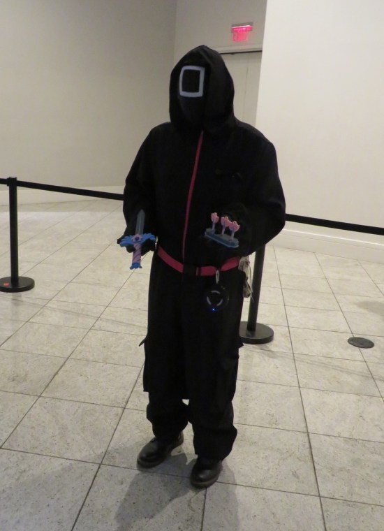 Squid Game black costume with square on mask, holding a dagger and darts, each pink and blue.