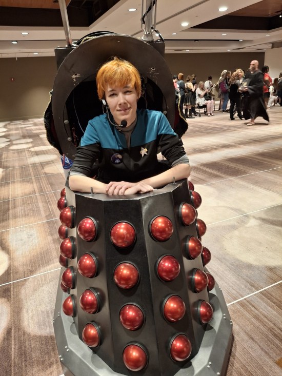 Trek fan in Voyager Science Officer top, seated inside a black Dalek covered in red half-spheres.