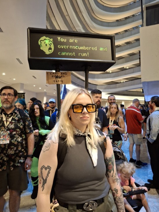 Woman in post-apocalyptic garb with backpack. Sticking out of it is a rectangular sign with Vault-Boy's panicky face saying "You are overencumbered and cannot run!"