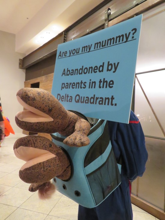 Trek cosplayer with backpack that has three stuffed brown salamanders sticking out of it, plus a sign reading "Are you my mummy? Abandoned by parents in the Delta Quadrant."