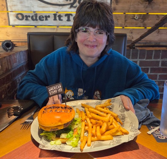 Anne smiling with a plate of burger and fries. Bun has Ford's logo burned into it and an "American Standard" toothpick flag.