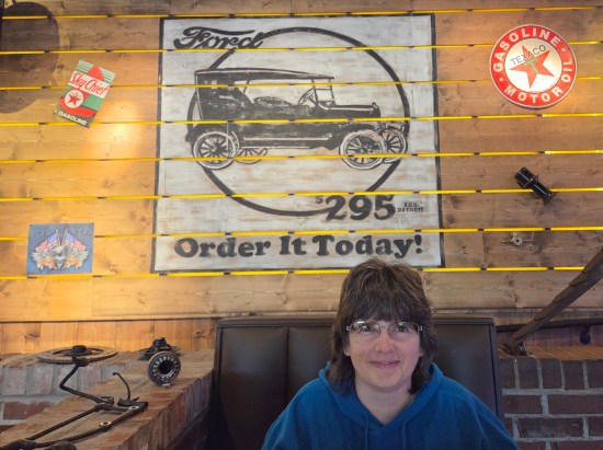 Anne sitting in front of a sign with a car on it reading "Ford $295 Order it today!" Wall is wood-paneled and has car-related mementos hanging on it.