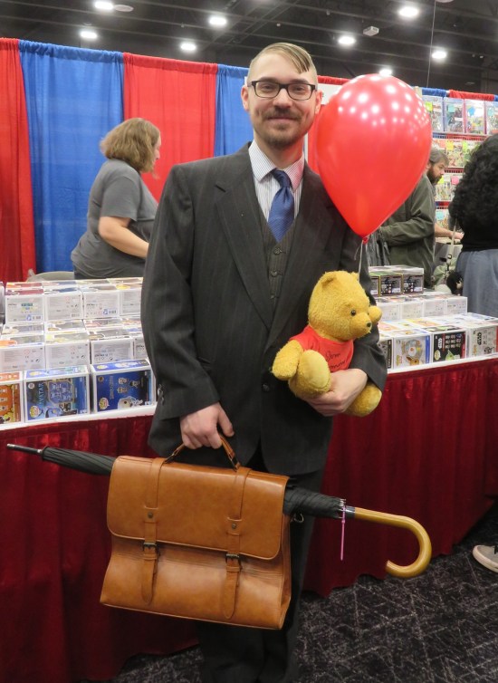 Christopher Robin cosplayer with suitcase, umbrella, stuffed Pooh and red balloon.