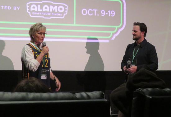 Two people in caption talking with mics in front of movie screen.