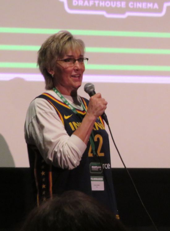 Christy Salters in an Indiana Pacers #22 jersey and white long-sleeved shirt, holding a mic in front of a movie screen.