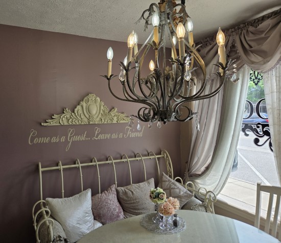 White table indoors near a window with an off-white metal outdoor sofa next to it, covered with four throw pillows. On the light purple wall is written, "Come as a guest...leave as a friend."