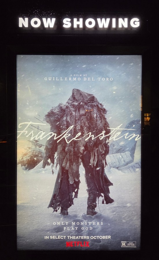 Frankenstein Movie Poster 2025 displayed outside a theater at night. The monster is a gangly tatterdemalion behind the logo and the logline "Only Monsters Play God".