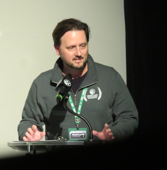 Greg Sorvig at a glass podium with big mic and wearing a gray Heartland hoodie.