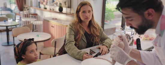 A middle-aged woman and a tiny girl at the counter of a brightly lit bakery, where the chef is icing a cake.