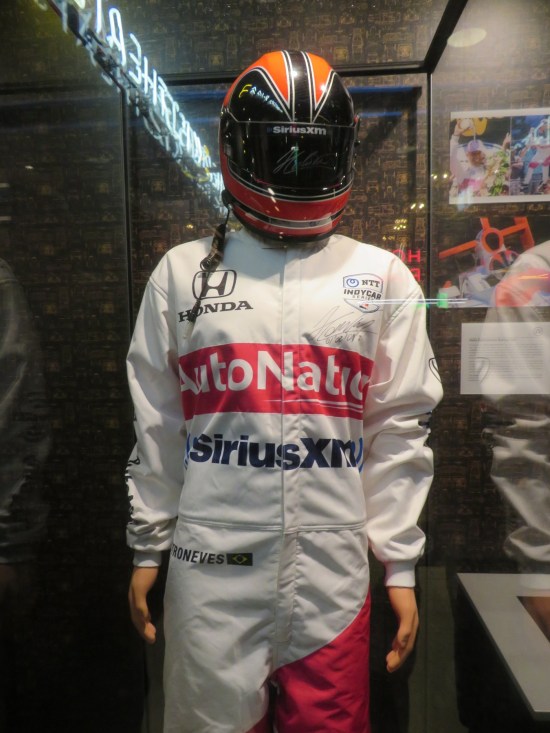 Race car driver jumpsuit and helmet in a vitrine with labels from Honda, AutoNation and SiriusXM.