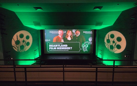 balcony view of a movie screen down below. Onscreen is a pic of Emily Deschanel and two producers, encouraging viewers to become a Heartland member. Walls around screen are glowing green and have large icons of film reels.