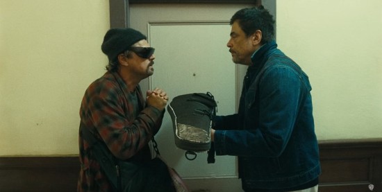 Benicio Del Toro hands a rifle in its storage bag to Leonardo DiCaprio, who looks like a frazzled mountain man with expensive sunglasses.