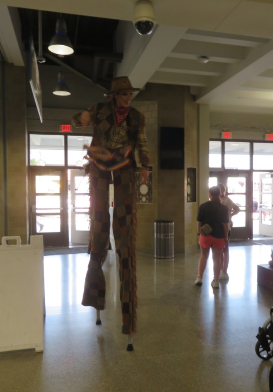 stilt-walker dressed like a cowpoke, walking indoors, slightly hunched and waving hi.
