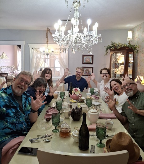 Seven people around a long tearoom table, all doing jazz hands, including this writer and his wife.