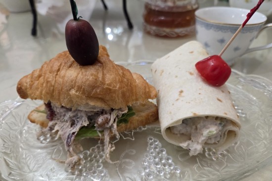 A tiny chicken salad sandwich topped with a kalamata olive on a toothpick, and a tiny deli wrap topped with a cherry tomato.