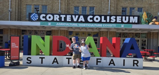 us doing jazz hands in front of a large Indiana State Fair logo standing outside the Corteva Coliseum.