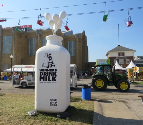 A giant milk bottle that says "Winners Drink Milk" sitting on grass next to a sidewalk. Behind it drives one of the fair's shuttle tractors, bea