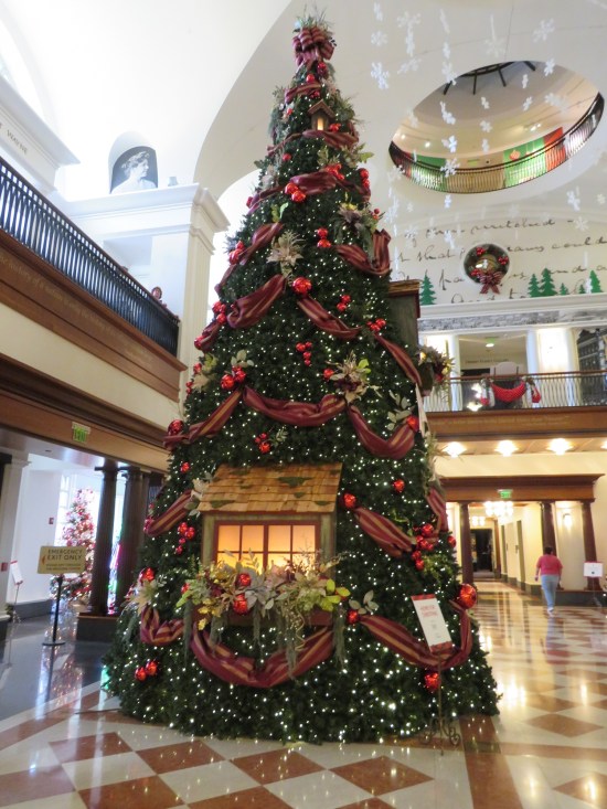 30-foot Christmas tree in the middle of a museum lobby, with red ribbons and a rustic cabin window.