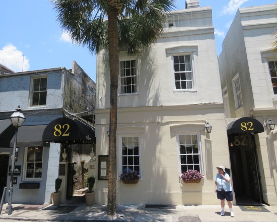 Two-story white brick building with black canopies on either side with gold '82' logos. Gated entrance is open at left, Anne approaches at right.