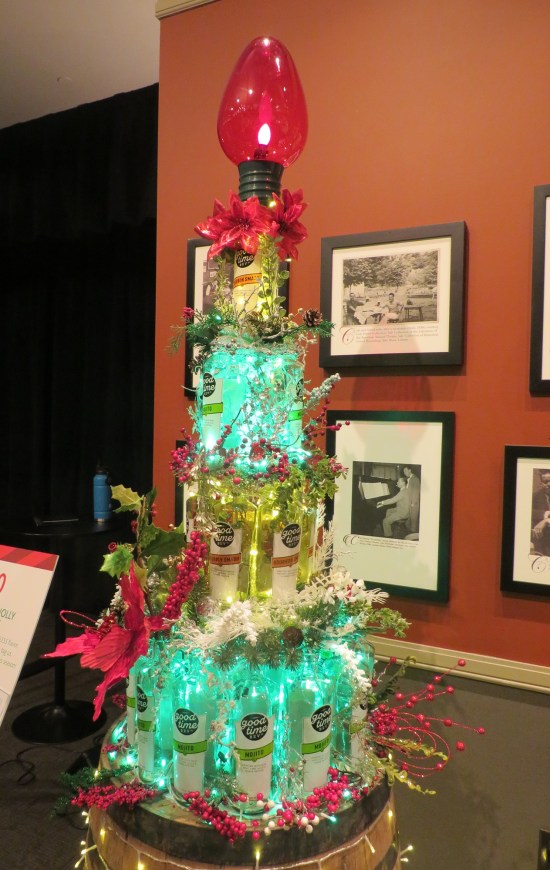 Bottles of liquor stacked in a tree shape on a table, topped with a single giant red Christmas light.