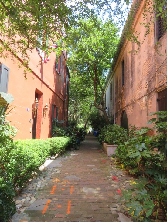 Sunny brick alley with trees in the distance and cultivated greenery lining either side of the peach buildings it runs between.