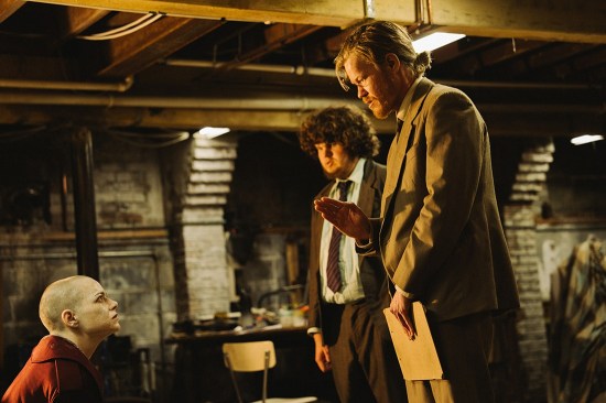 In a basement, Jesse Plemons and Aiden Delbis in rumpled suits standing over a seated and angry Emma Stone, whose head is shaven.