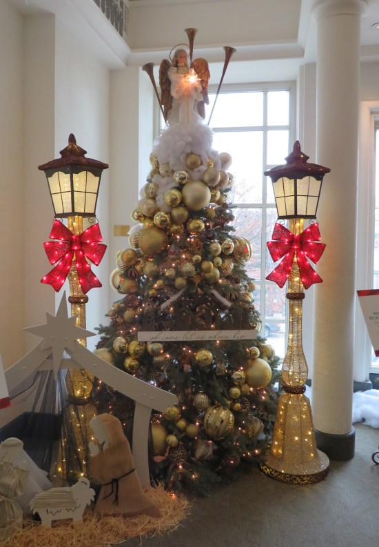 Christmas tree with large gold ball ornaments, huge angel topper with long heavenly trumpets. On each side is a fake streetlight with a big red bow. In front is a 2-D nativity scene.