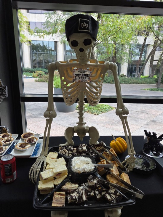 Table of party snacks presided over by a plastic skeleton with a chef's hate and nametag reading Chef Bones.