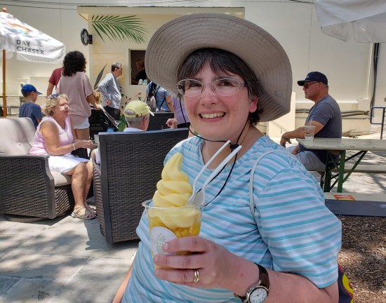 Anne in hat, smiling and holding yellow ice cream in cone. Pineapple Hut is in the far back. Customers sit outside in patio furniture.