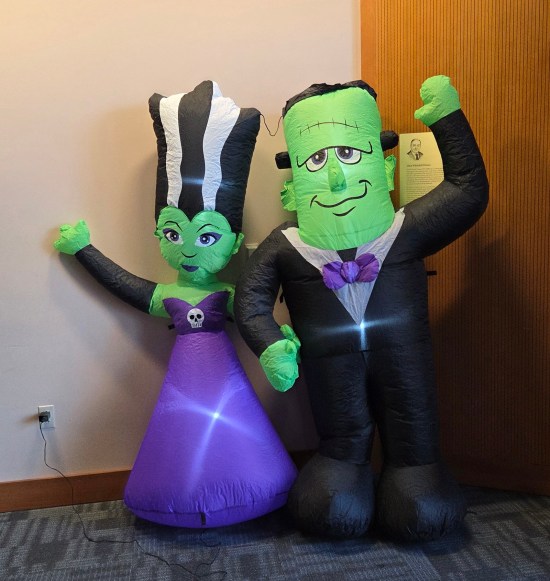 Inflatable Frankenstein monster and Bride of Frankenstein, each dressed nicely, holding hands and waving with their free hands, plugged in and standing in a corner.