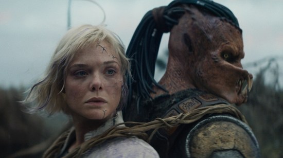 Elle Fanning and guy in Predator costume standing back to back, sternly.