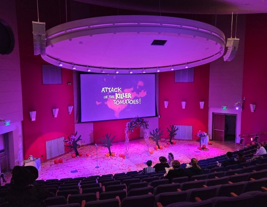 Auditorium stage with a bit of wedding decor, all swathed in purple lighting. On a large screen is the logo for "Attack of the Killer Tomatoes".