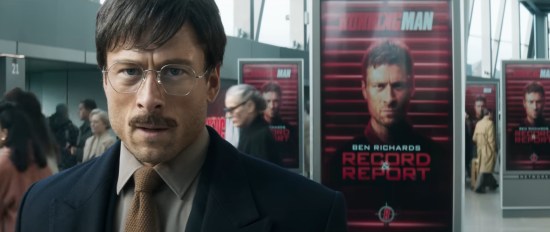 Glen powell disguised with glasses, mustache, and boring hair, very tense in a science fiction hallway filled with red WANTED posters for his character Ben Richards.