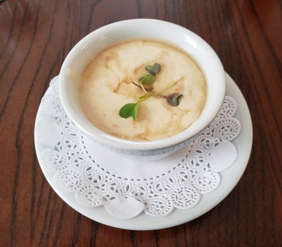 Cup of she-crab soup topped with a few herbs, served in white cup on white saucer with white doily on hardwood table.