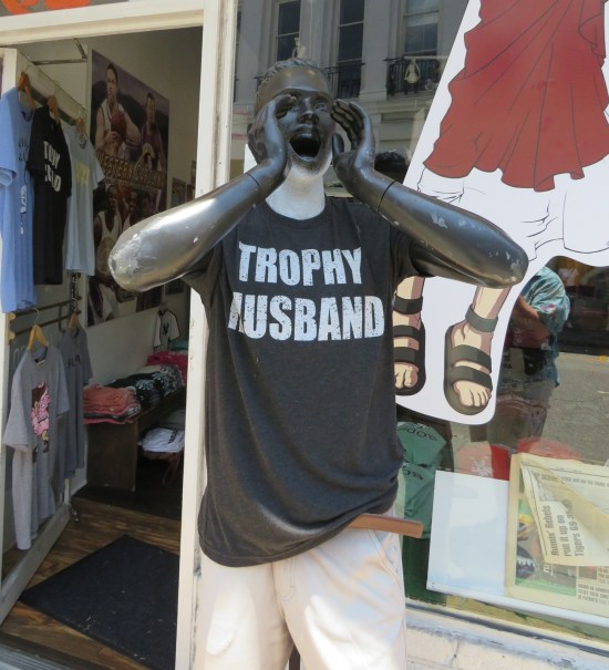 Gray mannequin posed outside a sports shop to appear as if screaming at passersby. Wearing a shirt that says "Trophy Husband".