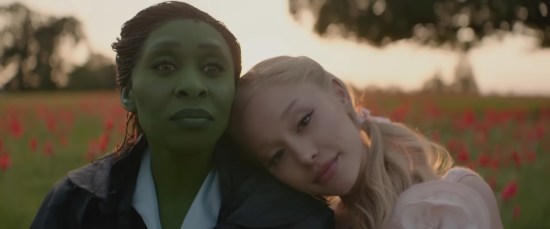 Glinda lays her head on Elphaba's shoulder as they sit smiling in a peaceful meadow.
