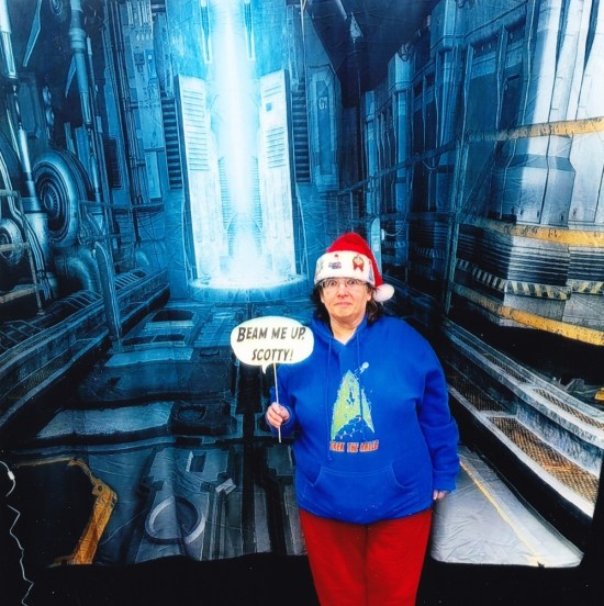 Anne holding a word balloon on a stick reading "BEAN ME UP, SCOTTY!" Behind her is a spaceship backdrop, a blue interior resembling a warp core or a Borg regeneration alcove, not sure what it actually is.