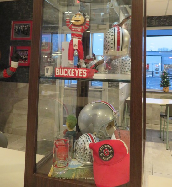 Another glass display with one Brutus Buckeye doll and several football helmets.