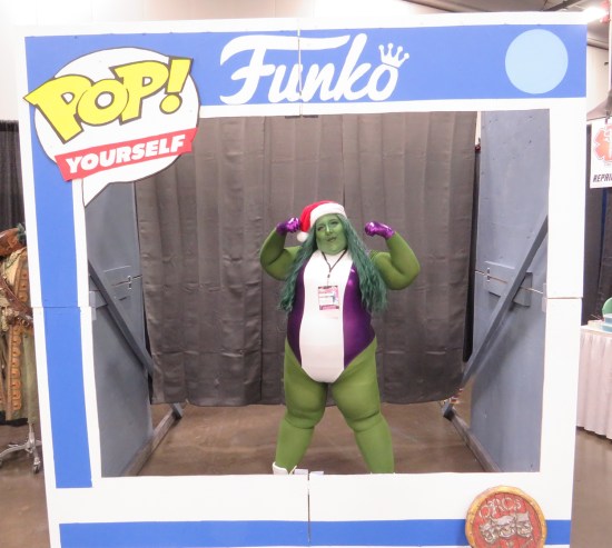 She-Hulk cosplayer with Santa cap standing inside a giant Funko Pop box, flexing her arms.