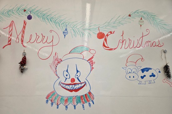 whiteboard with "Merry Christmas" written on it, plus marker drawings of a cow and an evil clown, both wearing Santa hats. Two decorated pinecones hang from magnets.