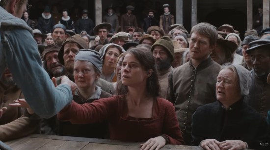 Jessie Buckley in the front row of a standing Shakespearean audience, reaching out to the actor on stage.