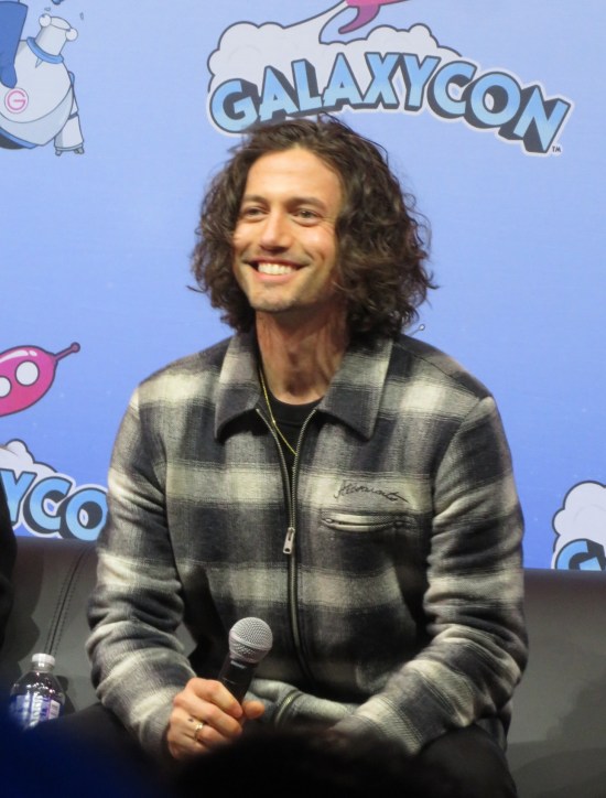 Jackson Rathbone in black-and-white flannel shirt, sitting on GalaxyCon stage and smiling at the audience.