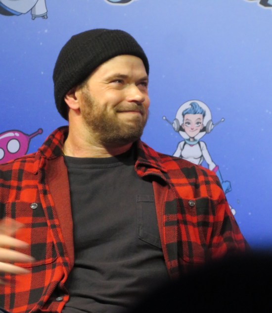 Kellan Lutz in red-and-black flannel and black lumberjack cap, sitting on stage and smiling.