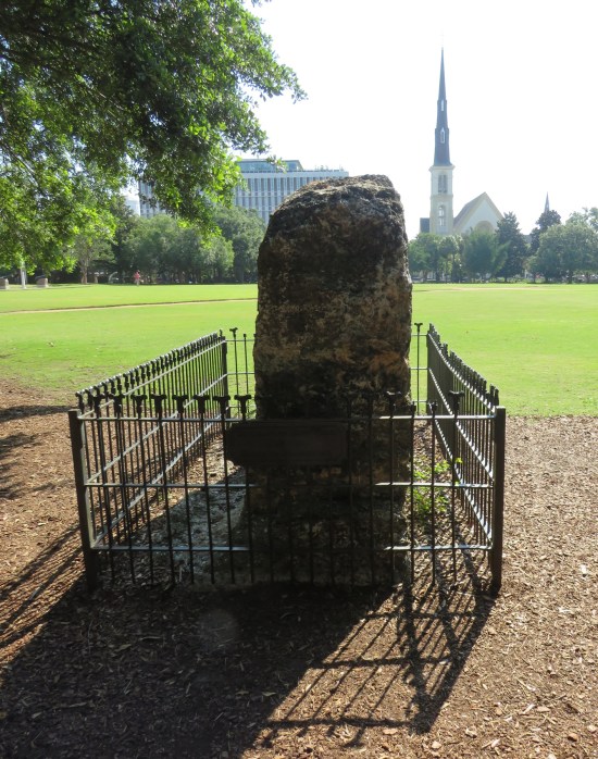 Short, lumpy stone column surrounded by tiny black metal fence, in the middle of a happy park.