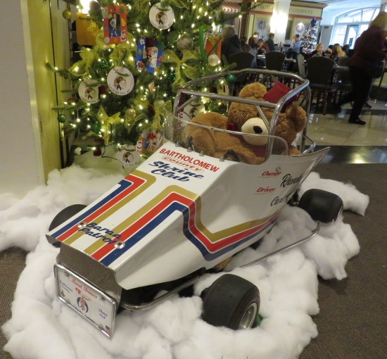 teddy bear in the driver seat of a tiny model Shriner car under a Christmas tree.