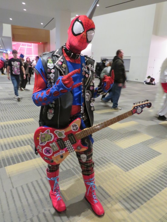 Spider-Punk costume with guitar, making devil-horns gesture.