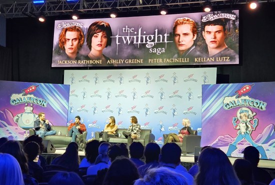 Slightly distant view of the four Twilight actors sitting on stage with a moderator. Above them is a digital banner with the series logo and their characters' head shots.